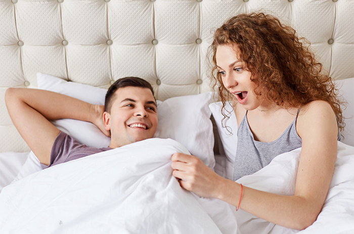 Shocked female looks under white blanket being surprised with size of boyfriends erection