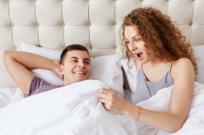 Shocked female looks under white blanket, being surprised with size of boyfriend`s penis after taking a Stamina for Men instant erection pill